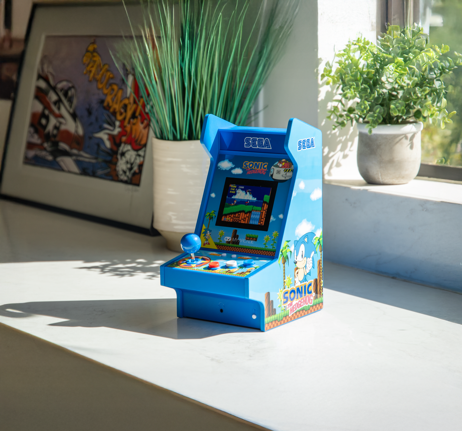 Sega Sonic-themed mini arcade machine on a table with plants and artwork in the background